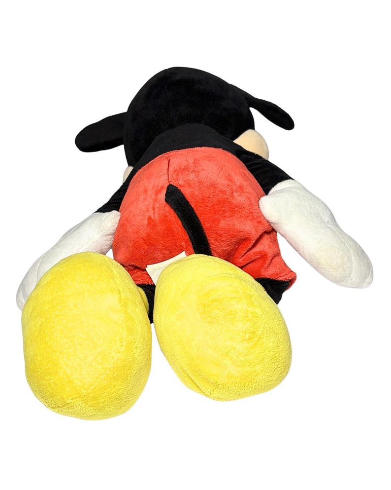 May include: A plush Mickey Mouse toy, viewed from the back. The toy features a black head with large ears, red shorts, yellow feet, and white gloves. The toy is soft and cuddly, perfect for children.