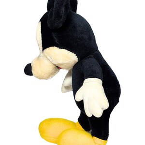 May include: A plush toy of a cartoon mouse, featuring a black body, ears, and nose, with a cream-colored face and hands. The mouse wears yellow shoes. The toy is posed in a side view against a white background.
