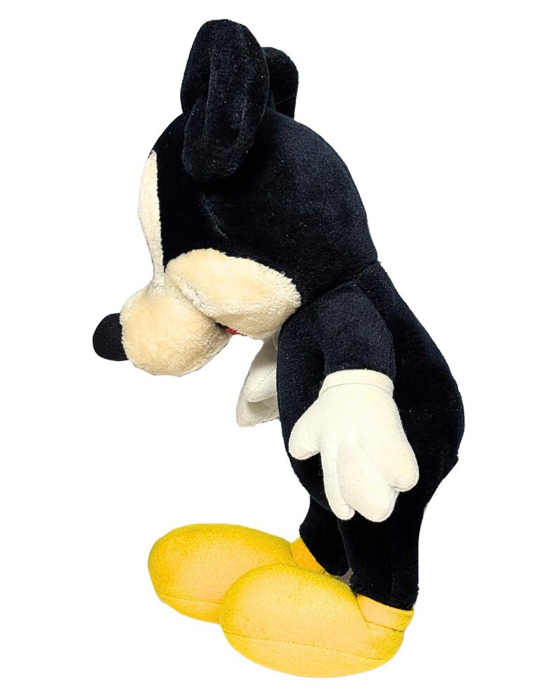 May include: A plush toy of a cartoon mouse, featuring a black body, ears, and nose, with a cream-colored face and hands. The mouse wears yellow shoes. The toy is posed in a side view against a white background.
