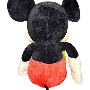May include: A plush Mickey Mouse toy, viewed from the back. The toy features black fur, large round ears, a red pair of shorts, and white gloves. The tail is black. The toy is a classic Disney character.