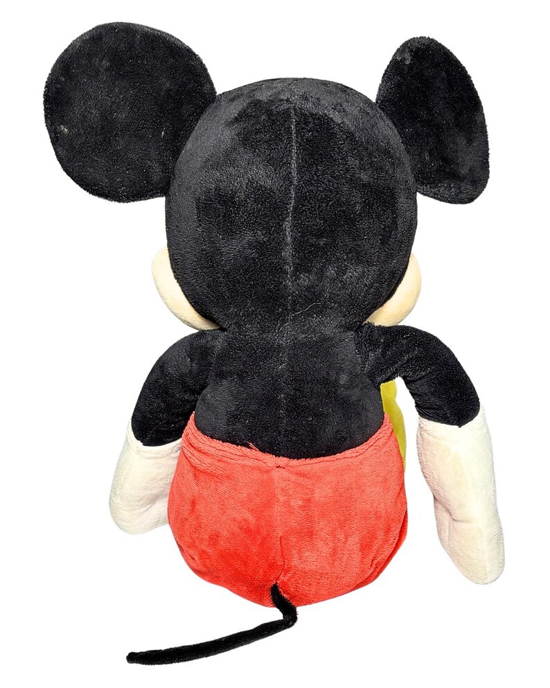 May include: A plush Mickey Mouse toy, viewed from the back. The toy features black fur, large round ears, a red pair of shorts, and white gloves. The tail is black. The toy is a classic Disney character.