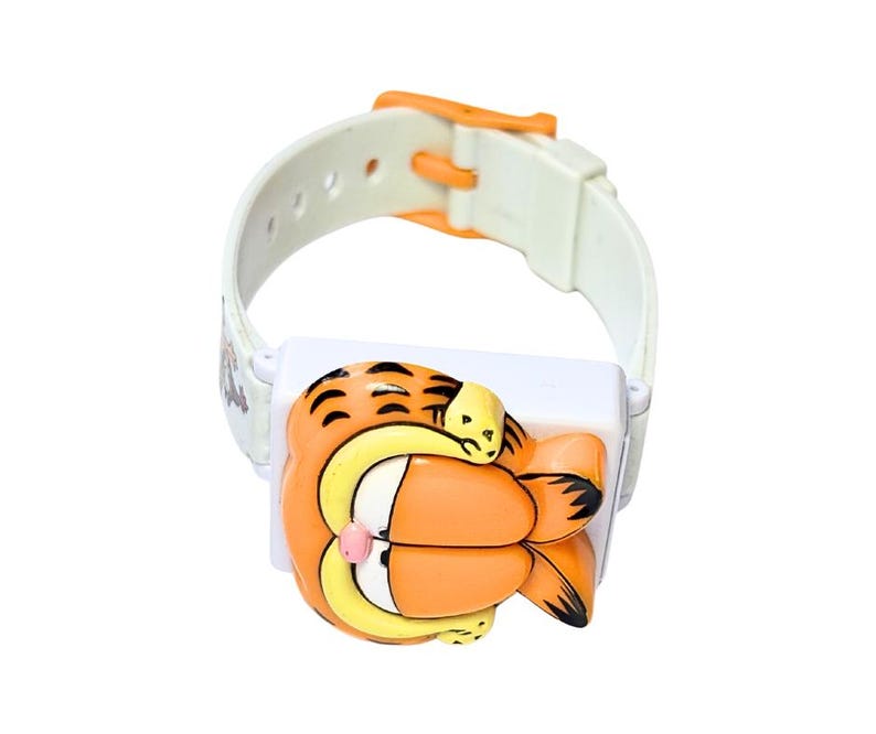 May include: A white and orange Garfield watch with a cartoon Garfield character design. The watch has a white strap with an orange buckle. The watch face features a 3D Garfield character in orange, yellow, and black.