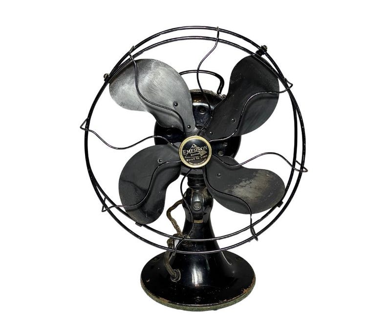 May include: Vintage Emerson electric fan with four blades and a protective wire cage. The fan is black with a circular base and a central emblem. The blades are a mix of black and gray. The fan is a classic piece of home decor.