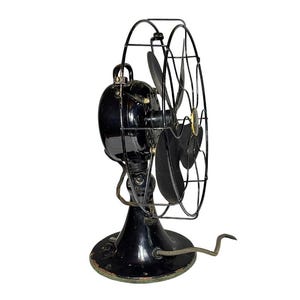 May include: An antique black electric fan with a cage and three blades. The fan has a round base and a cord. The fan is a vintage item, likely from the early 20th century. The fan's blades are black and the cage is made of metal.