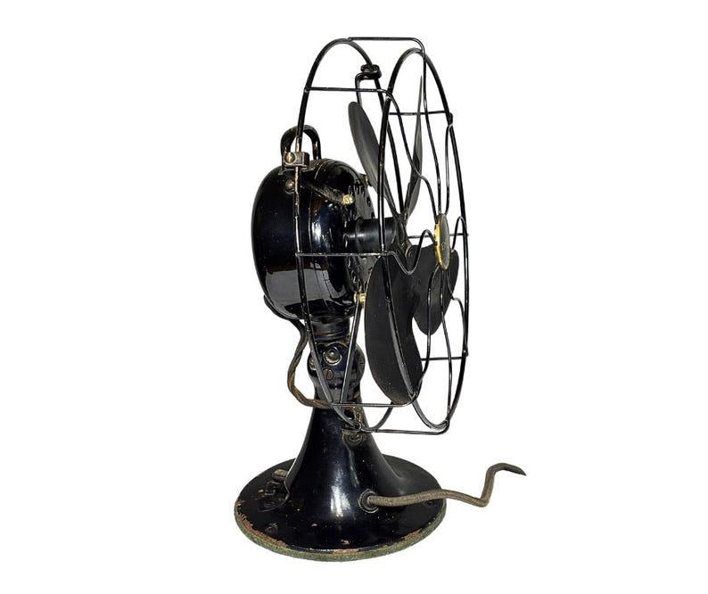 May include: An antique black electric fan with a cage and three blades. The fan has a round base and a cord. The fan is a vintage item, likely from the early 20th century. The fan's blades are black and the cage is made of metal.