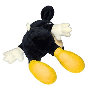 May include: A plush Mickey Mouse toy, lying on its stomach. The toy is primarily black with white gloves and a yellow pair of shoes. The ears are black and stand up. The toy is on a white background.