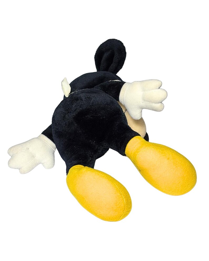 May include: A plush Mickey Mouse toy, lying on its stomach. The toy is primarily black with white gloves and a yellow pair of shoes. The ears are black and stand up. The toy is on a white background.