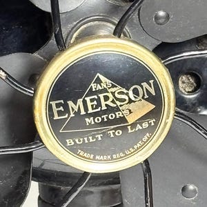May include: Close-up of an Emerson Motors fan. The fan's center features a gold-rimmed emblem with the words "EMERSON MOTORS" and "BUILT TO LAST" in white text on a black background. The fan blades and protective cage are visible.
