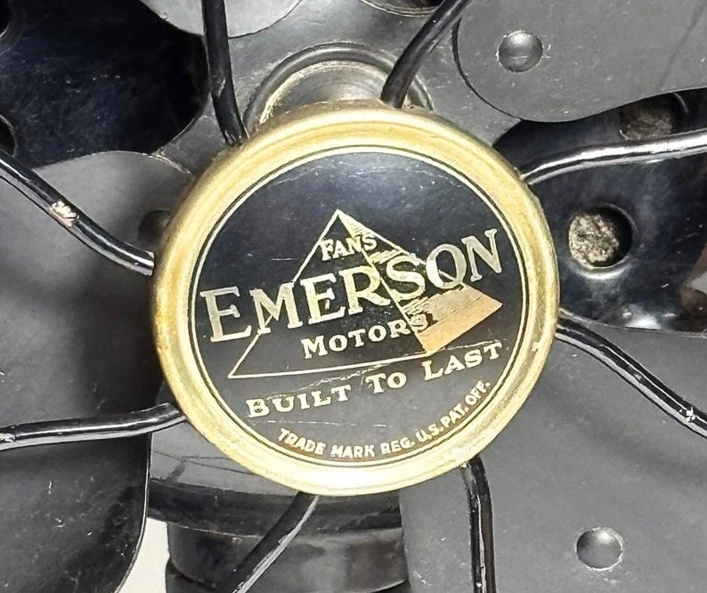 May include: Close-up of an Emerson Motors fan. The fan's center features a gold-rimmed emblem with the words "EMERSON MOTORS" and "BUILT TO LAST" in white text on a black background. The fan blades and protective cage are visible.