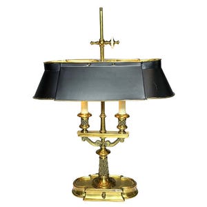 May include: A brass desk lamp featuring a rectangular black metal shade, two light sockets with white candle covers, and a central vertical post with an adjustable knob. The base is oval-shaped with a textured, cross-hatched pattern on the stem.