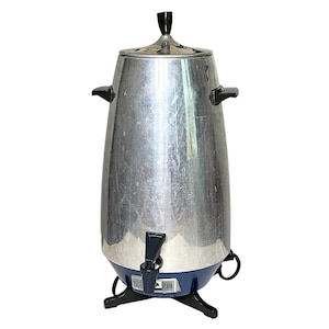 May include: A tall, metallic silver electric coffee percolator with a tapered body, black plastic side handles, a black lid knob, and a black spigot. The base is dark blue with a black plastic stand featuring four feet and a power cord attachment.