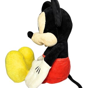 May include: A plush Mickey Mouse toy in a seated position. The toy features a black head and ears, a cream-colored face and hands, red shorts, and yellow feet. The toy has a black tail and is set against a white background.