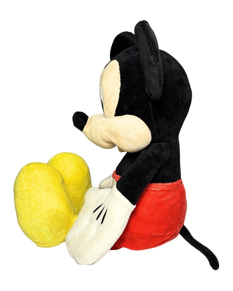 May include: A plush Mickey Mouse toy in a seated position. The toy features a black head and ears, a cream-colored face and hands, red shorts, and yellow feet. The toy has a black tail and is set against a white background.