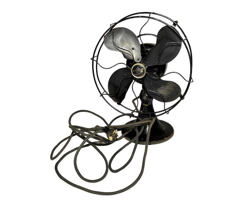 May include: An antique Emerson electric fan with a black metal frame and blades. The fan has a circular cage and a base, with a power cord extending from the bottom. The blades are black and silver.
