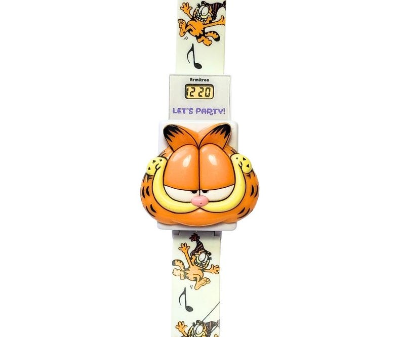 May include: A vintage Garfield watch with an orange Garfield head and a white band. The watch face displays the time as 12:20. The band features Garfield illustrations and musical notes. The text "LET'S PARTY!" is printed on the watch.