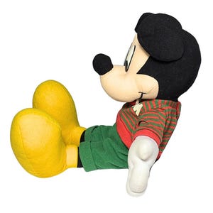 May include: A plush Mickey Mouse doll in a relaxed pose. Mickey is wearing a red and green striped sweater, green pants, and yellow shoes. His black ears and nose are prominent, and his white gloves are visible.