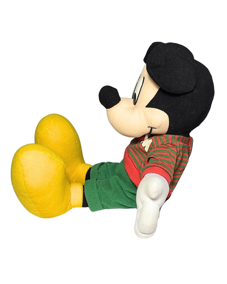 May include: A plush Mickey Mouse doll in a relaxed pose. Mickey is wearing a red and green striped sweater, green pants, and yellow shoes. His black ears and nose are prominent, and his white gloves are visible.