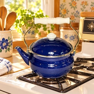 May include: A deep blue enamel tea kettle with a glossy finish, featuring a gold-toned metal handle with a white wooden grip, a matching white lid knob, and a curved spout, resting on a black metal stovetop grate in a kitchen setting.