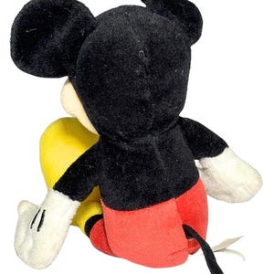 May include: A plush Mickey Mouse toy, viewed from the back. The iconic character features a black head and ears, a white face, red shorts, yellow feet, and white gloves. A black tail extends from the back. The toy is soft and cuddly.