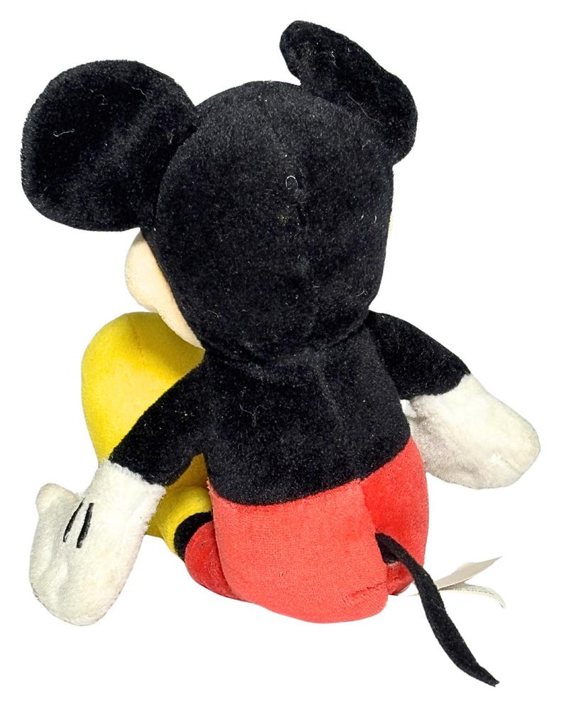 May include: A plush Mickey Mouse toy, viewed from the back. The iconic character features a black head and ears, a white face, red shorts, yellow feet, and white gloves. A black tail extends from the back. The toy is soft and cuddly.