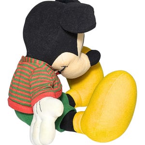 May include: A plush Mickey Mouse toy in a seated position. Mickey has a black head and ears, a red and green striped sweater, green pants, yellow shoes, and white gloves. The toy is set against a white background.