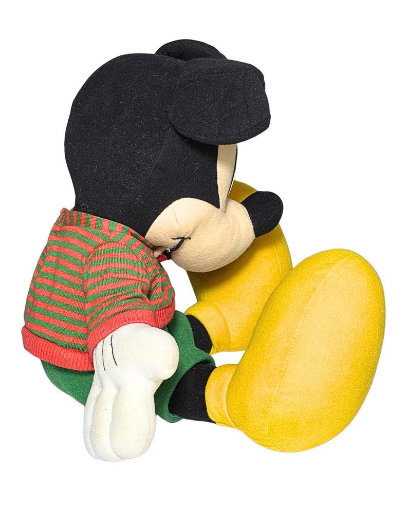 May include: A plush Mickey Mouse toy in a seated position. Mickey has a black head and ears, a red and green striped sweater, green pants, yellow shoes, and white gloves. The toy is set against a white background.
