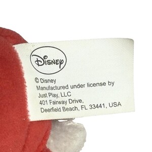 May include: A white tag with the Disney logo and text. The tag includes the text "&copy; Disney", "Manufactured under license by Just Play, LLC", and the address "401 Fairway Drive, Deerfield Beach, FL 33441, USA".