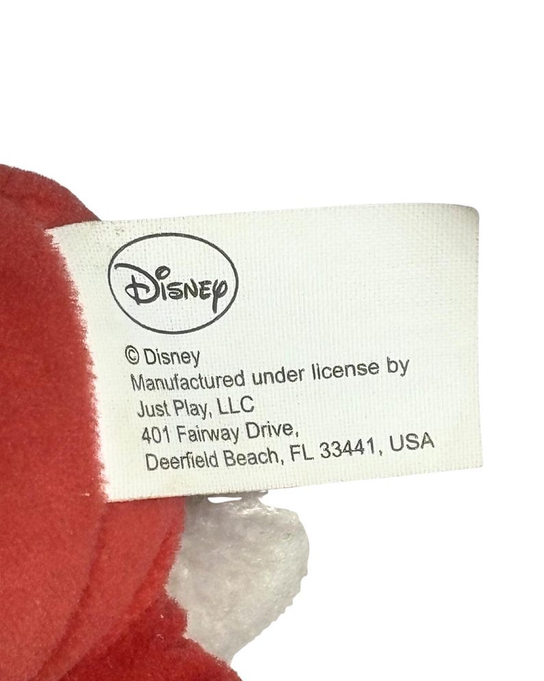 May include: A white tag with the Disney logo and text. The tag includes the text "&copy; Disney", "Manufactured under license by Just Play, LLC", and the address "401 Fairway Drive, Deerfield Beach, FL 33441, USA".
