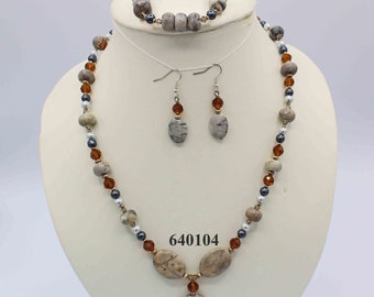 May include: A set of jewelry featuring a necklace, bracelet, and earrings. The necklace is made of brown, gray, and white beads with a pendant in the shape of a large oval. The bracelet is made of brown, gray, and white beads. The earrings are made of brown, gray, and white beads with a small oval pendant. The set is labeled with the number 640104.