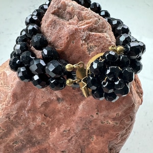 May include: A black beaded bracelet with a gold clasp. The bracelet is made of faceted black beads and is layered for a dramatic look.