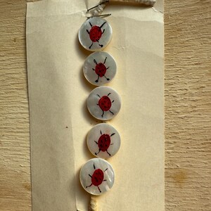 May include: Five round, white buttons with a red ladybug design, attached to a tan paper card. The ladybugs have black details. The buttons are held together with a metal pin and a piece of beige string.