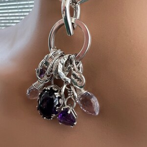 Puede incluir: Un collar de plata con un gran colgante circular y varios abalorios. Los abalorios incluyen piedras preciosas moradas y transparentes, y una piedra ovalada de color p&uacute;rpura oscuro. La cadena del collar est&aacute; hecha de eslabones de plata.