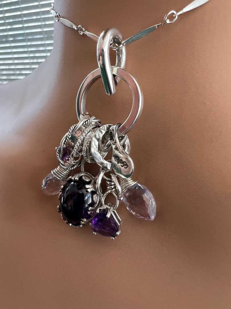 Puede incluir: Un collar de plata con un gran colgante circular y varios abalorios. Los abalorios incluyen piedras preciosas moradas y transparentes, y una piedra ovalada de color p&uacute;rpura oscuro. La cadena del collar est&aacute; hecha de eslabones de plata.