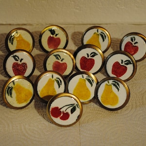 May include: A collection of decorative cabinet knobs with fruit designs. Each knob features a white background with a gold-colored rim and a painted image of a pear, apple, or cherries. The knobs are arranged on a white surface.