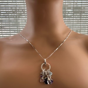 Puede incluir: Collar de plata con una cadena delicada y un colgante con un anillo que sostiene varios dijes de piedras preciosas en tonos morado, azul y rosa. El collar se exhibe en un maniqu&iacute;.