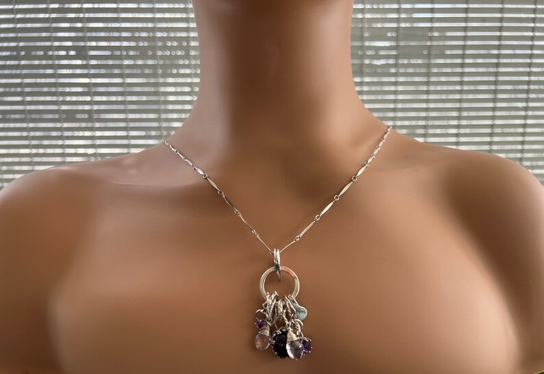 Puede incluir: Collar de plata con una cadena delicada y un colgante con un anillo que sostiene varios dijes de piedras preciosas en tonos morado, azul y rosa. El collar se exhibe en un maniqu&iacute;.