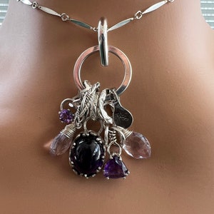 Puede incluir: Collar de plata con un gran colgante de anillo, adornado con varios abalorios y piedras preciosas. Los abalorios incluyen plumas, una herradura y una peque&ntilde;a etiqueta. Las piedras son amatista y cuarzo transparente.