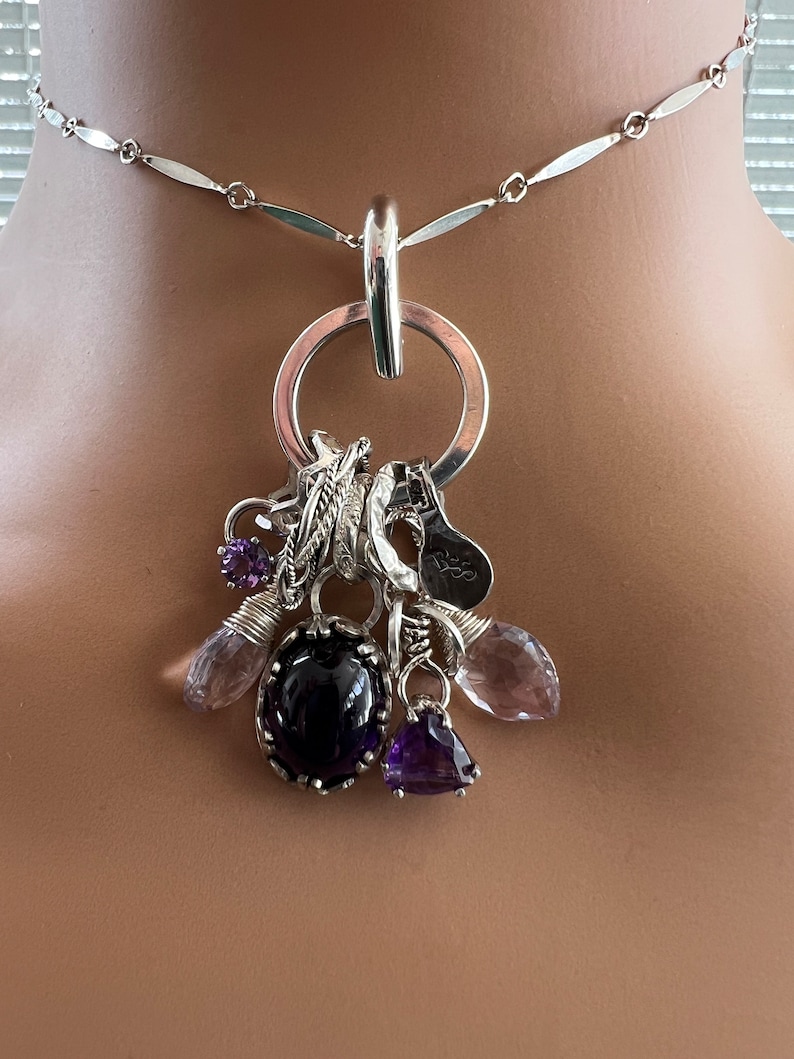 Puede incluir: Collar de plata con un gran colgante de anillo, adornado con varios abalorios y piedras preciosas. Los abalorios incluyen plumas, una herradura y una peque&ntilde;a etiqueta. Las piedras son amatista y cuarzo transparente.