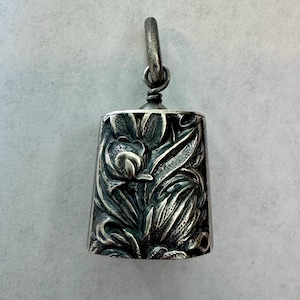 May include: A silver-toned metal pendant shaped like a bell, featuring a detailed embossed floral pattern with leaves and petals across its surface. The pendant includes a small, circular metal loop at the top for attachment to a chain or cord.