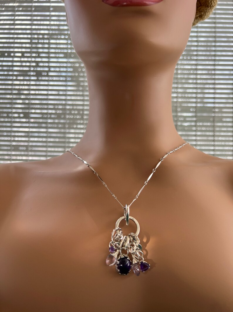 Puede incluir: Un collar de plata con un colgante con una gema grande de color p&uacute;rpura oscuro y gemas m&aacute;s peque&ntilde;as de color p&uacute;rpura y rosa. El colgante est&aacute; unido a una delicada cadena de plata.