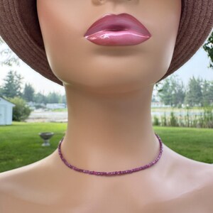 May include: A delicate, magenta-colored beaded choker necklace. The necklace is composed of small, evenly spaced beads, creating a subtle, elegant design. The necklace is displayed on a mannequin.