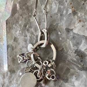 Puede incluir: Un collar de plata con un grupo de abalorios y piedras preciosas de amatista. El collar presenta un gran colgante circular y una cadena con eslabones en forma de barra. Los abalorios incluyen formas geom&eacute;tricas y un colgante redondo y en blanco.