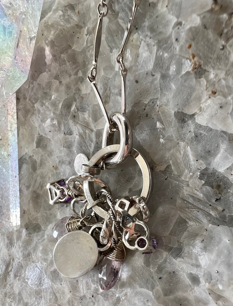 Puede incluir: Un collar de plata con un grupo de abalorios y piedras preciosas de amatista. El collar presenta un gran colgante circular y una cadena con eslabones en forma de barra. Los abalorios incluyen formas geom&eacute;tricas y un colgante redondo y en blanco.