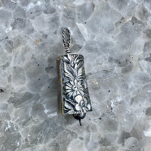 May include: A silver-toned bell pendant featuring an embossed floral pattern with detailed leaves and petals. The bell has a twisted metal loop at the top for attachment and a small clapper extending from the bottom, showcasing a metallic finish.