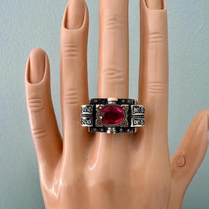 May include: A silver-toned ring with a large, oval, red gemstone at its center. The ring has an ornate design with small, clear accent stones and decorative patterns. The ring is displayed on a mannequin hand.