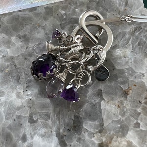 Puede incluir: Un collar de plata con m&uacute;ltiples colgantes, incluyendo piedras preciosas de amatista y peque&ntilde;os anillos. El collar presenta un dise&ntilde;o de bucle abierto grande y una cadena delicada. Las piedras preciosas son de varias formas y tonos de p&uacute;rpura.