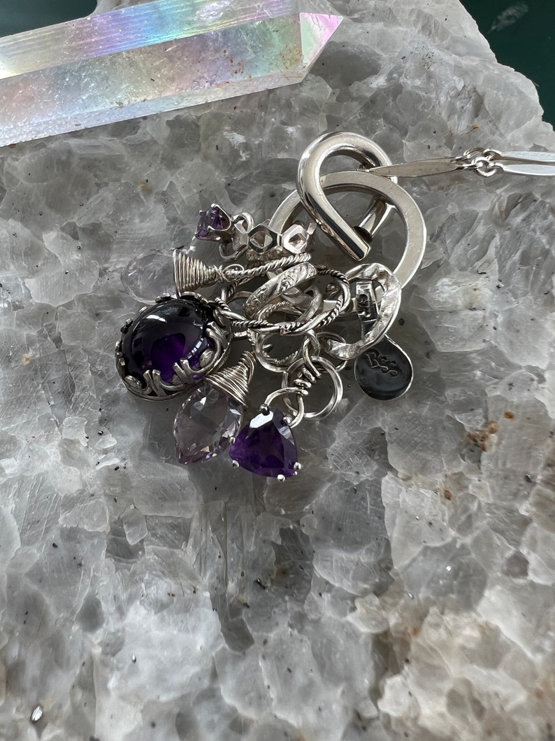 Puede incluir: Un collar de plata con m&uacute;ltiples colgantes, incluyendo piedras preciosas de amatista y peque&ntilde;os anillos. El collar presenta un dise&ntilde;o de bucle abierto grande y una cadena delicada. Las piedras preciosas son de varias formas y tonos de p&uacute;rpura.