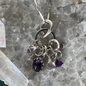 Puede incluir: Un collar con colgante de plata con un grupo de dijes y piedras preciosas. El collar incluye una gran fianza plateada curvada, m&uacute;ltiples anillos plateados y varios dijes. Los dijes incluyen amatistas y piedras preciosas de cuarzo transparente.