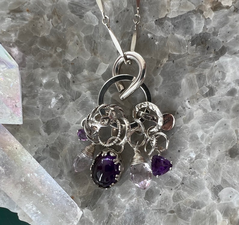 Puede incluir: Un collar con colgante de plata con un grupo de dijes y piedras preciosas. El collar incluye una gran fianza plateada curvada, m&uacute;ltiples anillos plateados y varios dijes. Los dijes incluyen amatistas y piedras preciosas de cuarzo transparente.