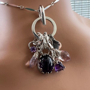 Puede incluir: Collar de plata con un gran colgante circular y un conjunto de abalorios. Los abalorios incluyen varias piedras preciosas moradas y peque&ntilde;os adornos plateados. El collar tiene una cadena delicada con eslabones alargados.